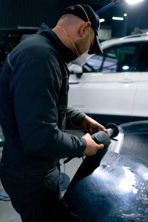 Téléchargez les photos : Dans une station-service, un mécanicien de voiture masqué essaie de poncer une partie de la voiture. - en image libre de droit