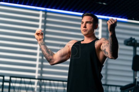 Foto de En el gimnasio joven chico guapo con tatuajes en una camiseta negra está haciendo un calentamiento en frente de la máquina de ejercicios - Imagen libre de derechos