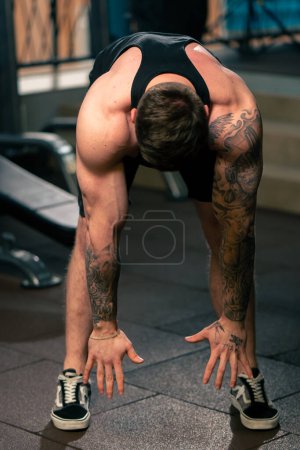 Téléchargez les photos : Dans la salle de gym jeune beau gars avec des tatouages dans un t-shirt noir fait un échauffement devant la machine d'exercice - en image libre de droit
