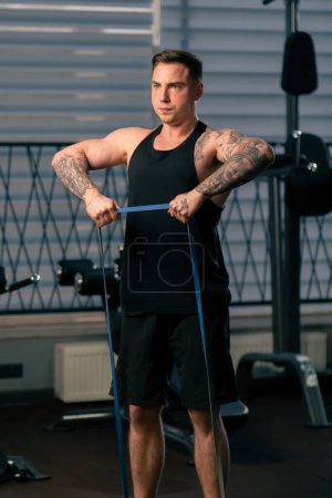 Foto de En el gimnasio un chico guapo joven con tatuajes en una camiseta negra hace ejercicios con una banda elástica - Imagen libre de derechos