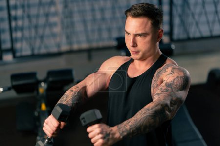 Foto de En el gimnasio un chico guapo joven con tatuajes en una camiseta negra hace ejercicios con dos pesas - Imagen libre de derechos
