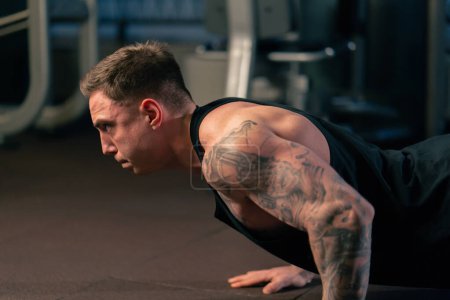 Photo for Close up in the gym a young handsome guy with tattoos in a black T-shirt does push-ups on the floor - Royalty Free Image