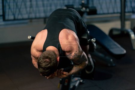 Photo for In the gym a young handsome guy with tattoos in a black T-shirt shakes his back on an exercise machine - Royalty Free Image