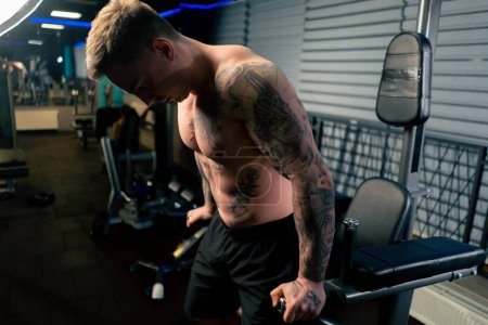 Téléchargez les photos : Dans la salle de gym jeune beau gars avec des tatouages se tire vers le haut et pompe ses bras sur la barre horizontale - en image libre de droit