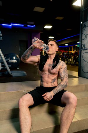 Téléchargez les photos : Dans la salle de gym jeune beau gars avec des tatouages assis boire de l'eau à partir d'une bouteille en plastique au repos - en image libre de droit