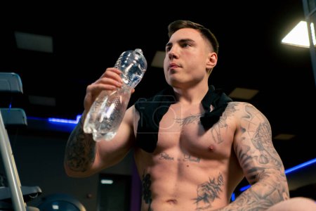Foto de Primer plano en el gimnasio joven guapo chico con tatuajes sentado beber agua de una botella de plástico descansando - Imagen libre de derechos