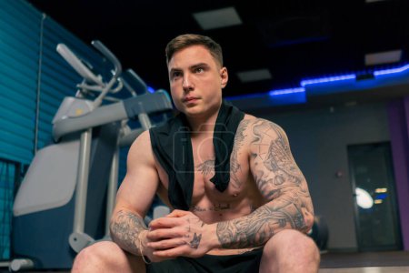 Téléchargez les photos : Dans la salle de gym un jeune beau gars avec des tatouages est assis au repos pense à un exercice avec les mains pliées - en image libre de droit