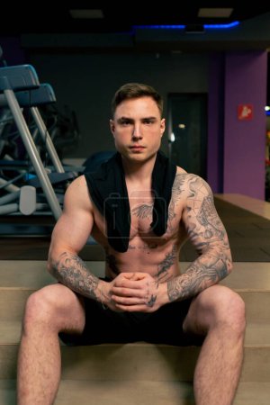 Photo for In the gym a young handsome guy with tattoos sits resting thinks about an exercise with his hands folded - Royalty Free Image
