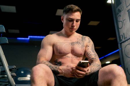 Téléchargez les photos : Dans la salle de gym un jeune beau mec avec des tatouages s'assoit et lit un message au téléphone - en image libre de droit