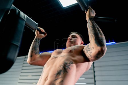Photo for Low shot in the gym a young handsome guy with tattoos is doing pull-ups on the horizontal bar shaking his back and arms - Royalty Free Image