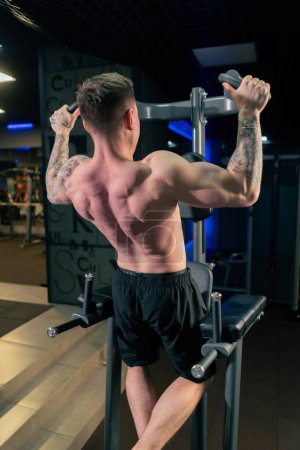 Foto de In the gym a young handsome guy with tattoos is doing pull-ups on the horizontal bar shaking his back and arms - Imagen libre de derechos