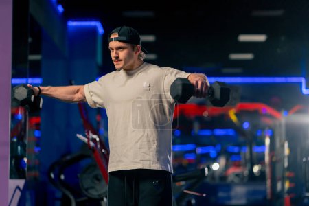 Photo for In the gym a young handsome guy in a T-shirt and a cap does exercises with dumbbells and shakes his arms - Royalty Free Image