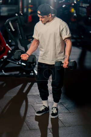 Photo for In the gym a young handsome guy in a T-shirt and a cap does exercises with dumbbells and shakes his arms - Royalty Free Image
