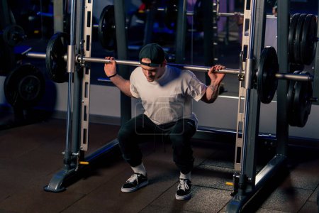 Photo for In the gym a young handsome guy in a T-shirt and a cap shakes his back with a barbell with athletic plates - Royalty Free Image