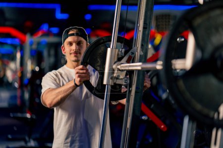 Photo for In the gym a young handsome guy in a T-shirt and a cap sets athletic plates on the barbell looks at the camera smiling - Royalty Free Image