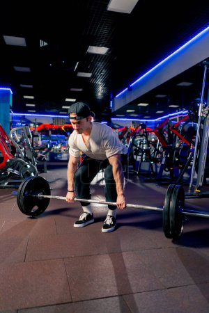 Photo for In the gym a young handsome guy in a T-shirt and a cap with a belt lifts a barbell with large athletic pancakes - Royalty Free Image