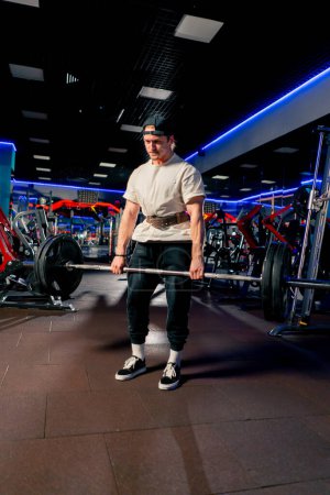 Foto de En el gimnasio un chico guapo joven en una camiseta y una gorra con un cinturón levanta una barra con grandes tortitas atléticas - Imagen libre de derechos