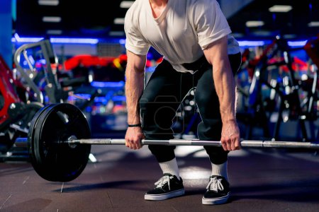 Foto de De cerca en el gimnasio un chico guapo joven en una camiseta y una gorra con un cinturón levanta una barra con grandes tortitas atléticas - Imagen libre de derechos