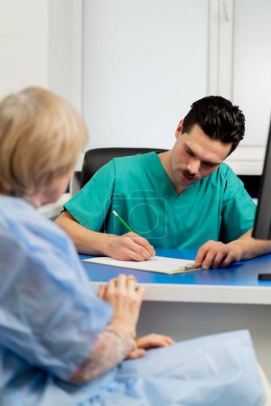 Téléchargez les photos : Dans une clinique de chirurgie plastique, un médecin consulte une femme âgée en prenant des notes sur papier - en image libre de droit