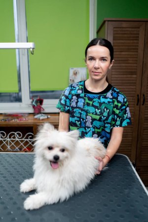 Foto de En un salón de aseo un peluquero con uniforme verde sostiene a un perro acostado en una mesa de aseo - Imagen libre de derechos