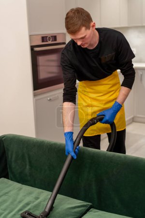 Foto de En un apartamento un limpiador en un delantal amarillo profesionalmente aspira un sofá de terciopelo verde con equipo profesional - Imagen libre de derechos