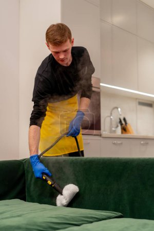 Téléchargez les photos : Dans un appartement un nettoyeur dans un tablier jaune fait un nettoyage à la vapeur d'un canapé vert avec un équipement professionnel - en image libre de droit