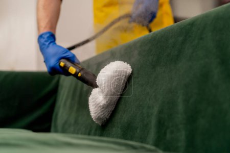 Téléchargez les photos : Fermer dans un appartement nettoyeur en tablier jaune fait un nettoyage à la vapeur d'un canapé vert avec un équipement professionnel - en image libre de droit