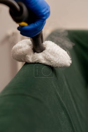 Foto de Primer plano en un limpiador de apartamentos en delantal amarillo hace una limpieza a vapor de un sofá verde con equipo profesional - Imagen libre de derechos