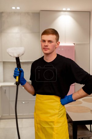 Photo for In an apartment cleaner in a yellow apron stands with a steam generator in his hand looking at the camera - Royalty Free Image