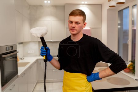Foto de En un limpiador de apartamentos en un delantal amarillo está parado con un generador de vapor en la mano mirando a la cámara - Imagen libre de derechos