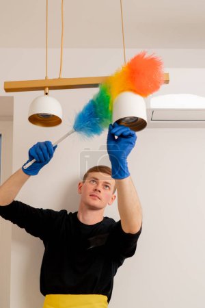 Photo for In the apartment a cleaner in a yellow apron is cleaning a chandelier with a dust brush - Royalty Free Image