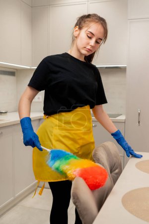 Foto de En el apartamento una señora de la limpieza en un delantal amarillo está limpiando sillas con un cepillo de polvo - Imagen libre de derechos