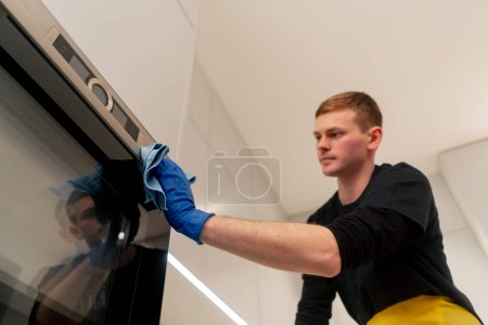 Foto de Tiro bajo en un apartamento un limpiador en un delantal amarillo está limpiando el horno con un trapo azul - Imagen libre de derechos
