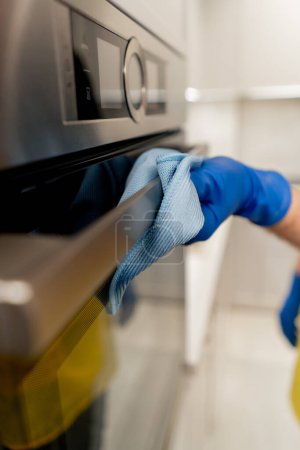 Photo for Close up in an apartment a cleaner in a yellow apron is cleaning the oven with a blue rag - Royalty Free Image