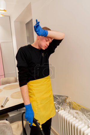Téléchargez les photos : In the apartment cleaner in yellow apron is tired wiping his forehead from sweat standing near the battery - en image libre de droit