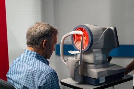 Foto de En una clínica de oftalmología un paciente de edad avanzada se sienta frente a una máquina de diagnóstico con una luz roja - Imagen libre de derechos