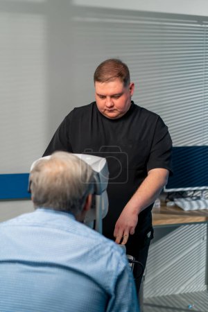 Foto de En una clínica de oftalmología, el médico obeso realiza un diagnóstico en un paciente de edad avanzada. - Imagen libre de derechos