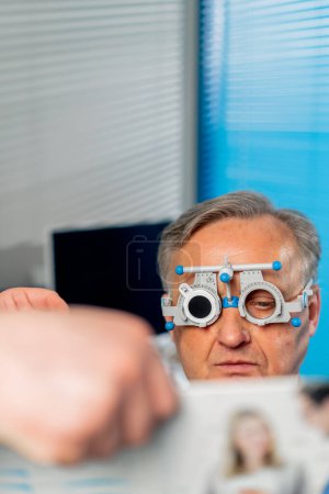 Téléchargez les photos : Gros plan dans une clinique d'ophtalmologie patient âgé sélectionne une lentille vérifie la vision en lisant - en image libre de droit