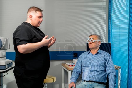 Foto de En una clínica de oftalmología un médico obeso consulta a un paciente de edad avanzada sobre lentes - Imagen libre de derechos