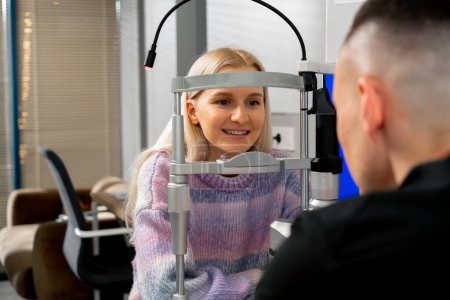 Foto de En una clínica de oftalmología una joven se comunica con un médico y sonríe a través de una máquina de diagnóstico - Imagen libre de derechos