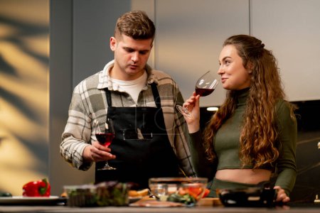 Foto de Pareja joven enamorada en hermosa cocina preparando la cena juntos y bebiendo vino - Imagen libre de derechos
