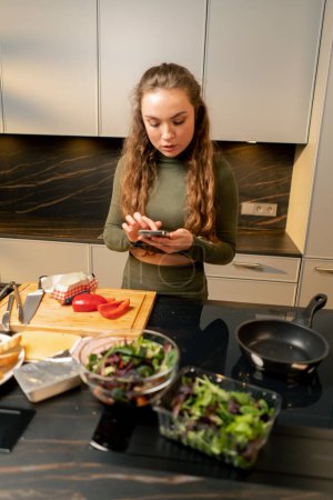 Téléchargez les photos : Jeune fille amoureuse dans une robe verte dans la cuisine préparer le dîner selon une recette de son téléphone - en image libre de droit