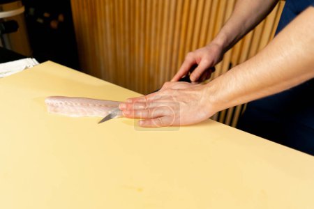 Téléchargez les photos : Gros plan dans un restaurant japonais un chef dans un uniforme bleu coupe le poisson pour les sushis en tranches sur un carton jaune - en image libre de droit