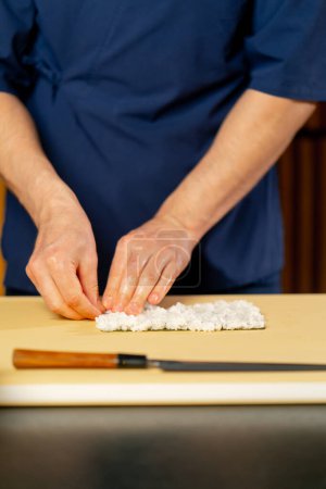 Téléchargez les photos : Gros plan dans un restaurant japonais chef en uniforme bleu sur un carton jaune tourne un rouleau de poisson - en image libre de droit