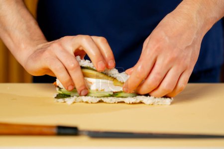 Téléchargez les photos : Gros plan dans un restaurant japonais chef en uniforme bleu sur un carton jaune tourne un rouleau de poisson - en image libre de droit