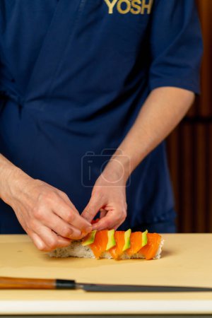 Foto de Primer plano en un chef de restaurante japonés en un uniforme azul en una tabla amarilla gira un rollo de pescado - Imagen libre de derechos