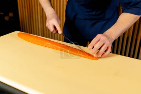 Foto de Primer plano en un restaurante japonés un chef en un uniforme azul corta pescado para sushi en rodajas en una tabla amarilla - Imagen libre de derechos