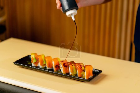 Foto de Primer plano en un restaurante japonés un chef en un uniforme azul vierte salsa sobre sushi preparado en un plato - Imagen libre de derechos