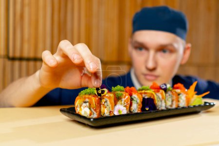 Téléchargez les photos : Gros plan dans un restaurant japonais chef en uniforme bleu décore des sushis prêts à l'emploi sur une assiette - en image libre de droit