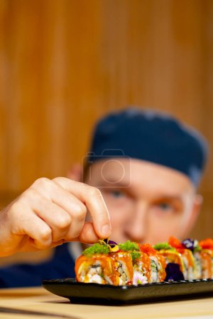 Téléchargez les photos : Gros plan dans un restaurant japonais chef en uniforme bleu décore des sushis prêts à l'emploi sur une assiette - en image libre de droit